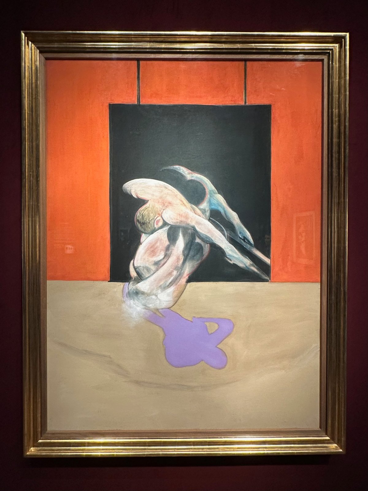 A twisting nude figure in a dark doorway against an orange wall, with a purple shadow on the floor, in a gilt frame